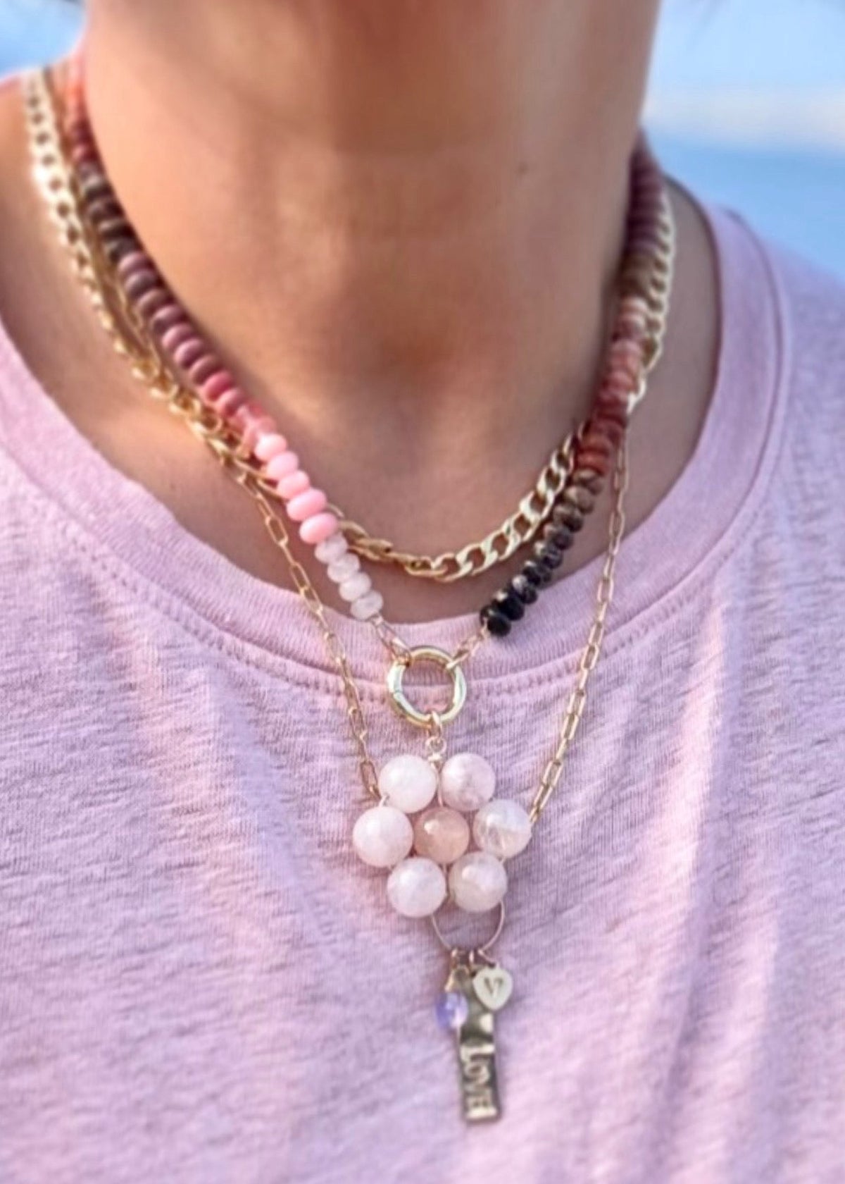 Necklace stack with a pink gemstone flower pendant, a cluster of charms, and a chunky gold chain on a light pink shirt